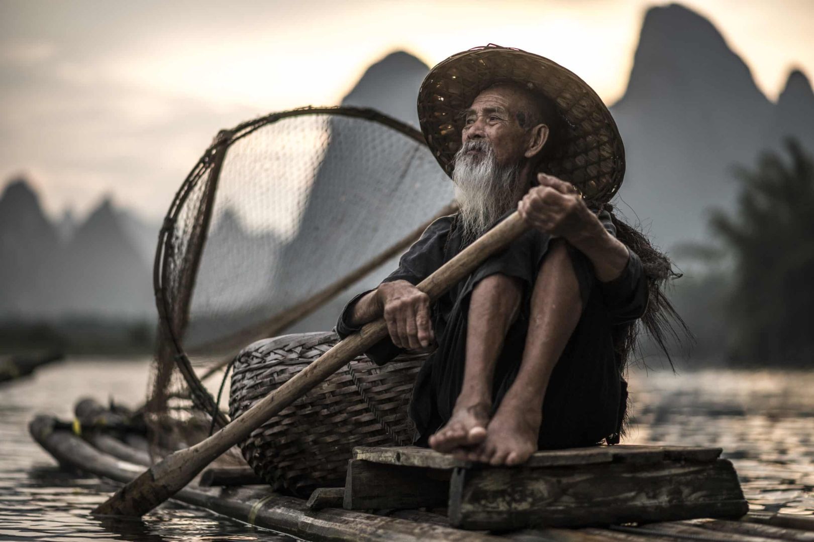 fotografía artística de retrato de un pescador anciano con cormoranes y sombrero sobre barca del río en china con barba y sujetando un remo